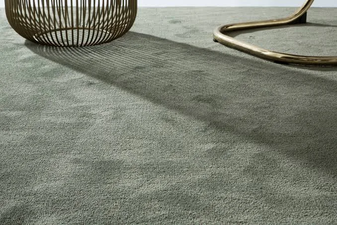 Moqueta de pelo corto verde salvia con luz rasante de tarde, bajo una mesa auxiliar de alambre y una silla de tubo de latón
